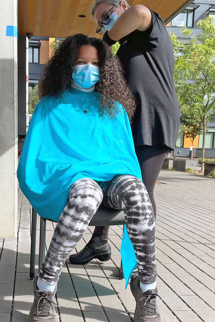 A girl with black curly hair wears a blue hairstyling cape and a face mask. A stylist, also wearing a face mask, cuts her hair
