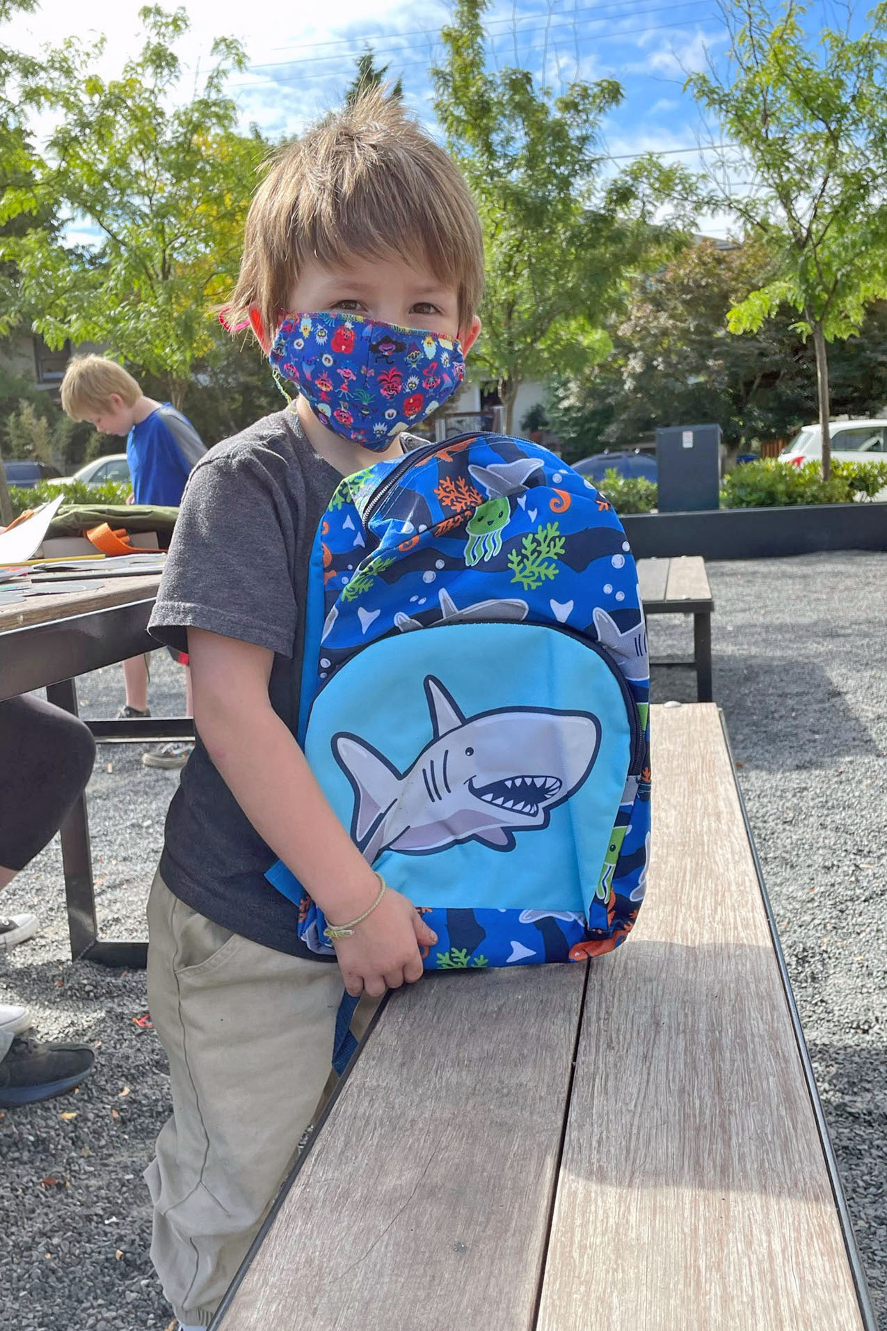 A young boy wears a face mask and holds up a backpack with a cartoon shark