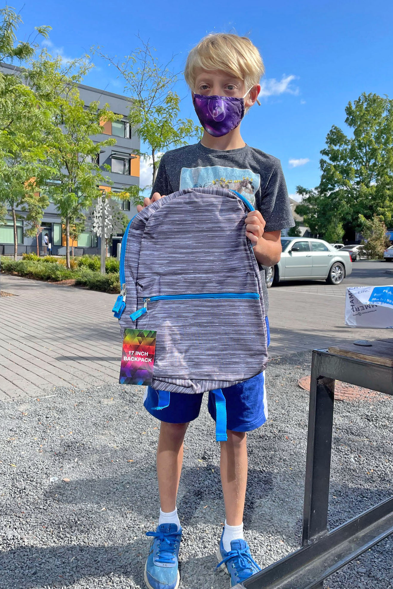 A boy wears a face mask, stands in front of a blue sky, and holds up a gray striped backpack