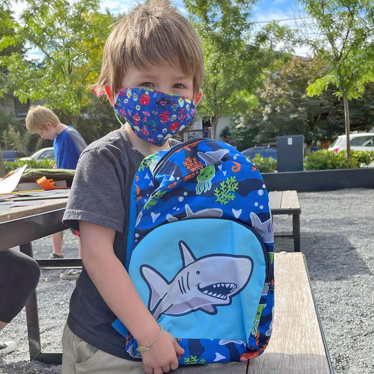 A young boy wears a face mask and holds up a kids' backpack with a cartoon shark
